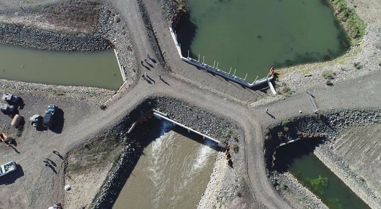 Aerial view looking straight down on gravel roads and parking areas, with several channels of water coming together, and water flowing through a white tide gate in the middle.